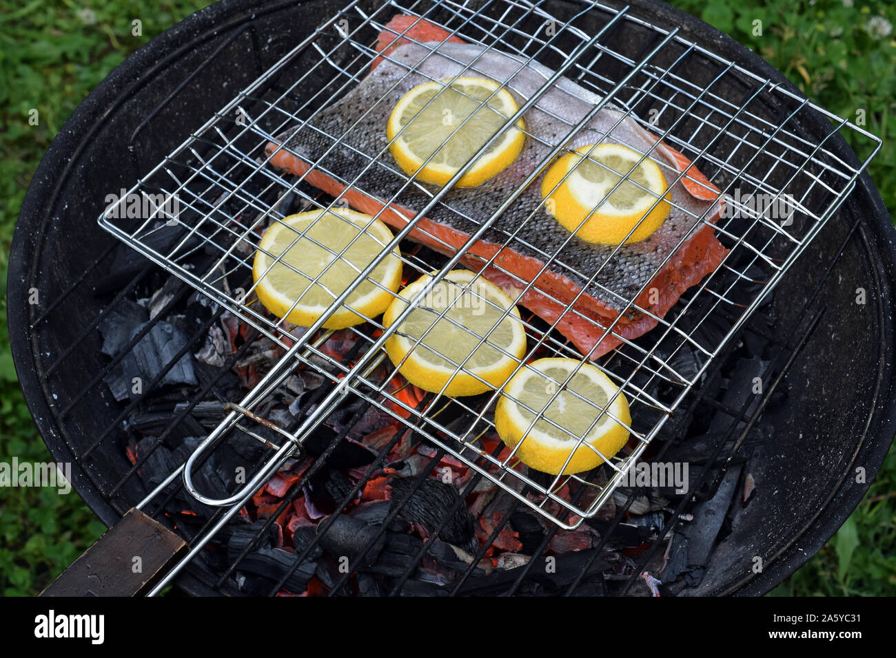 Grilling salmon with lemon slices on round charcoal grill with gridiron. Hot glowing embers under fish. Stock Photo