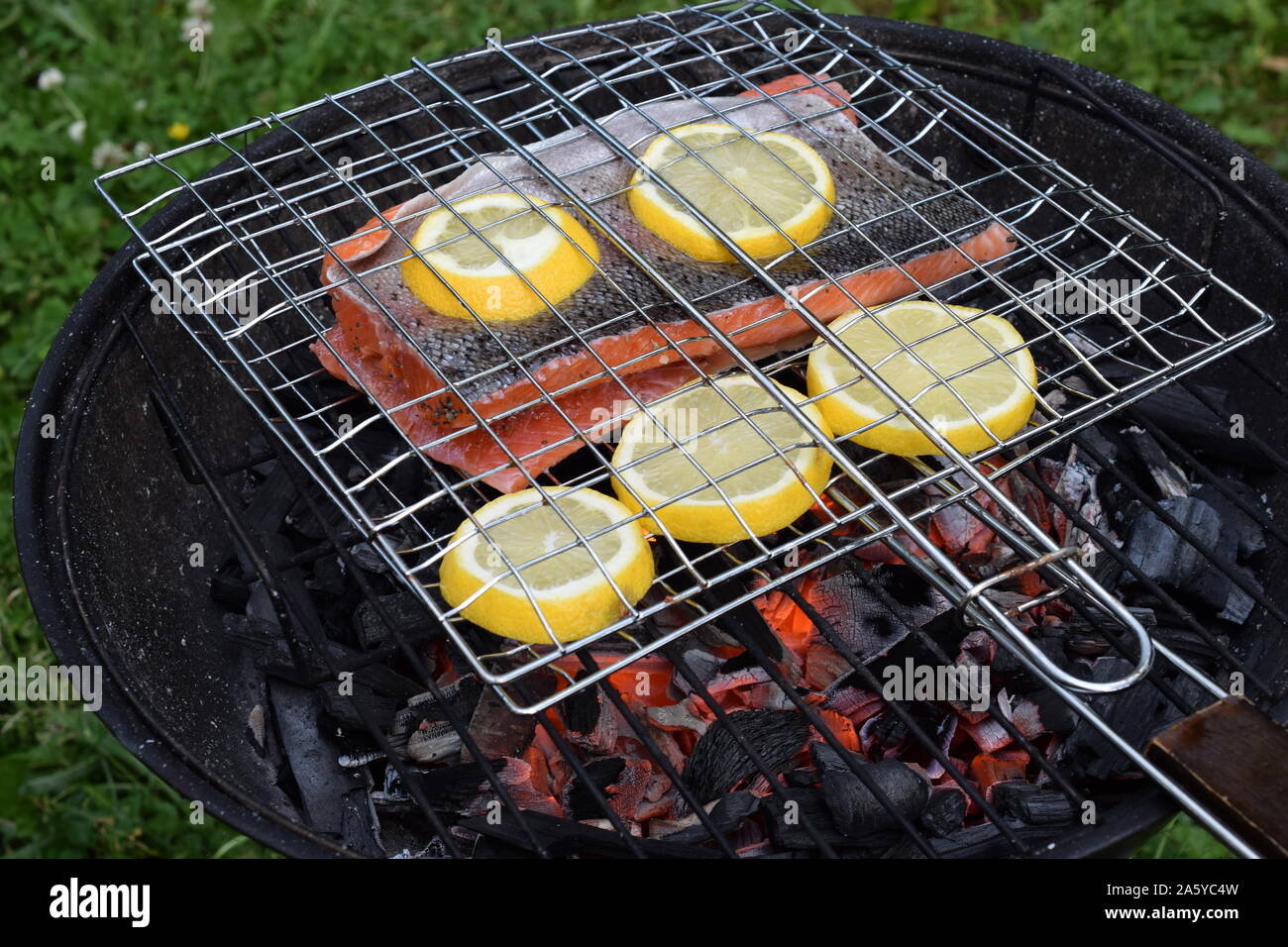 Grilling salmon with lemon slices on round charcoal grill with gridiron. Hot glowing embers under fish. Stock Photo