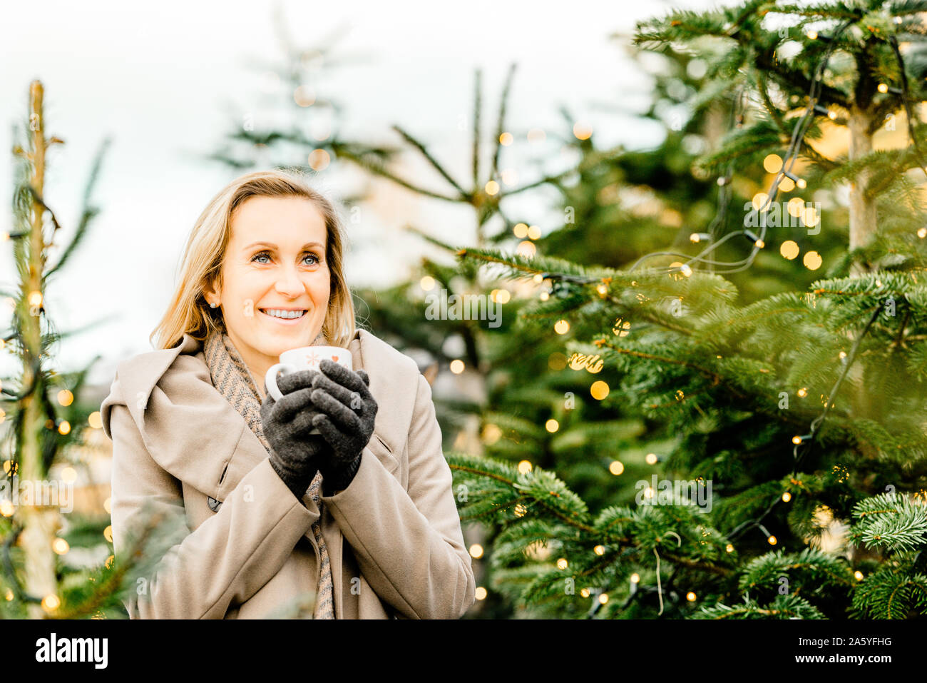portrait of a happy woman holding a hot drink in her hand surrounded by christmas lights on trees Stock Photo