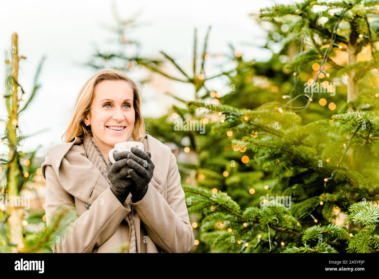 portrait of a happy woman holding a hot drink in her hand surrounded by christmas lights on trees Stock Photo