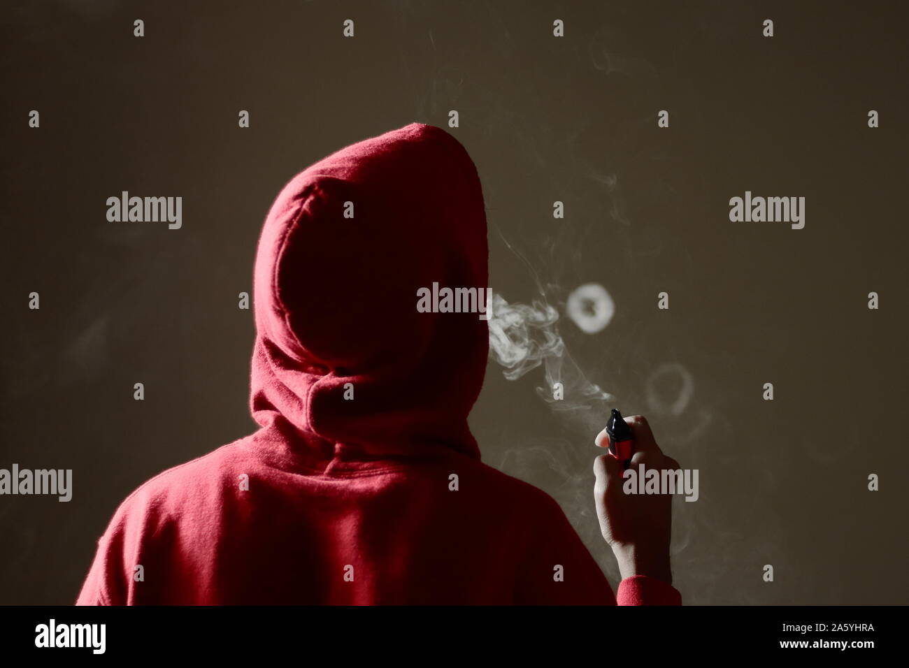 Young male in red hoodie vaping smoking, blows a single smoke ring while holding an electronic cigarette in hand, isolated rear view Stock Photo