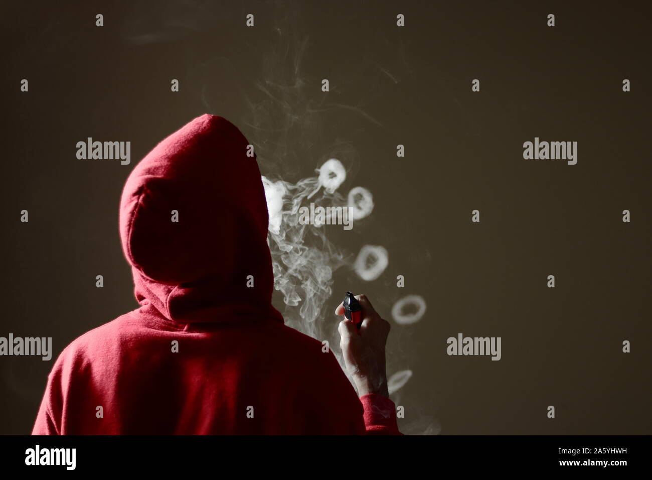 Young male in red hoodie vaping smoking, blows multiple smoke rings while holding a vape in hand, isolated rear view Stock Photo