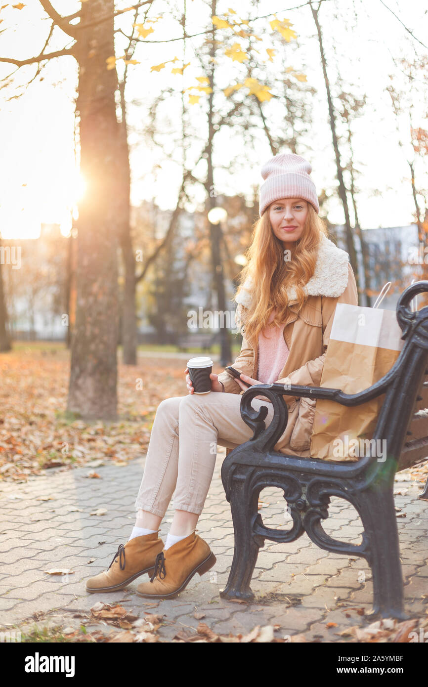 Redhead girl in warm clothes sitting with coffee on wooden bench in autumn park. Stock Photo