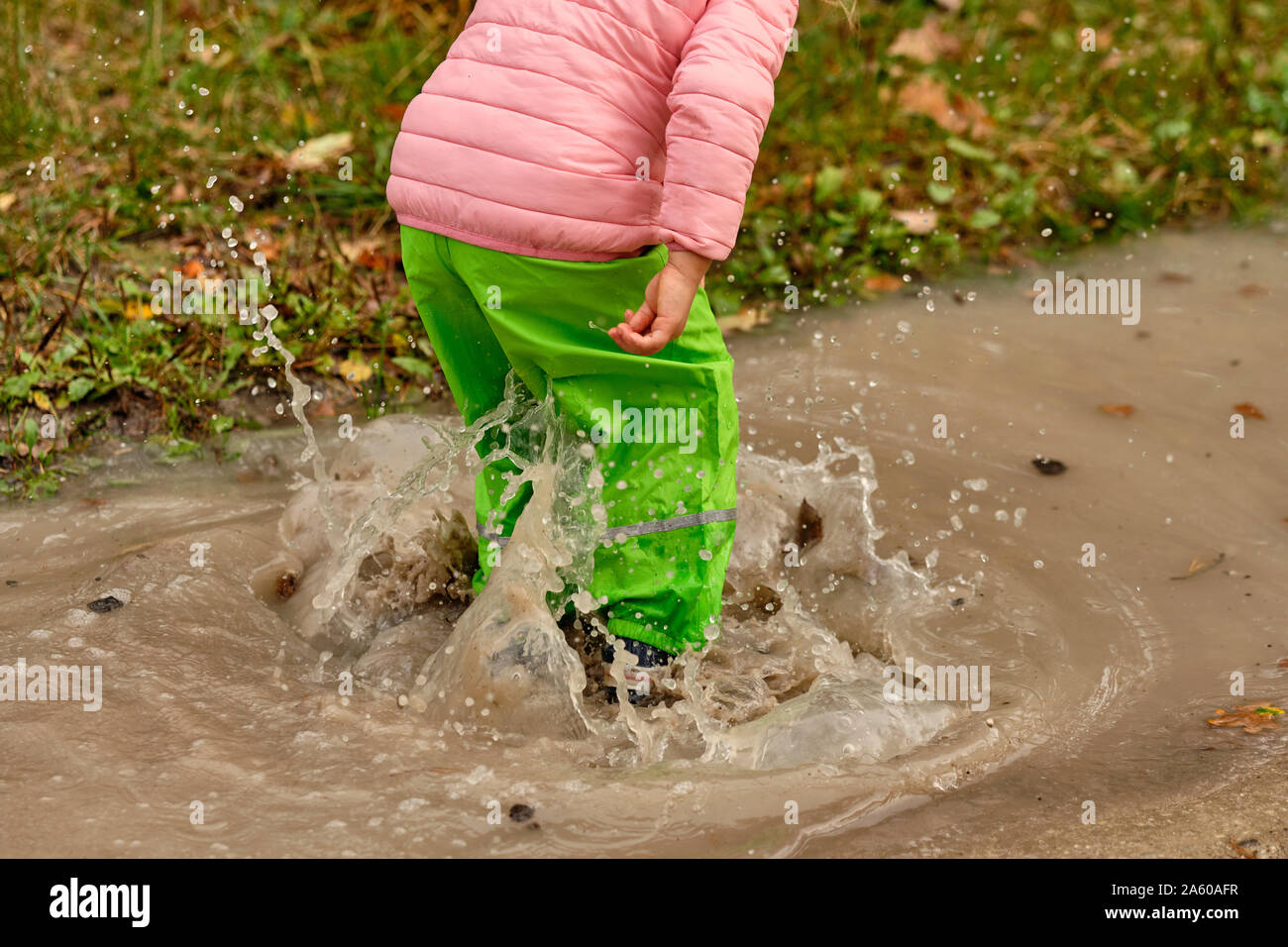 Low section of a child girl in green waterproof pants and rubber boots jumping into a massive rain puddle with the water splashing high up in the air Stock Photo