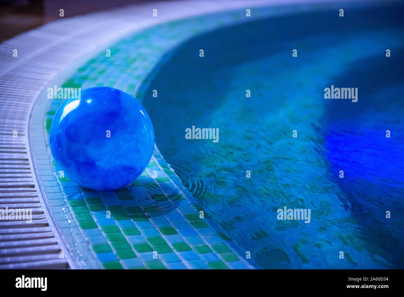 Blue ball is left in swimming pool decorated with blue and green mosaic tiles. Stairs are visible through shallow water. Summer season and private saf Stock Photo