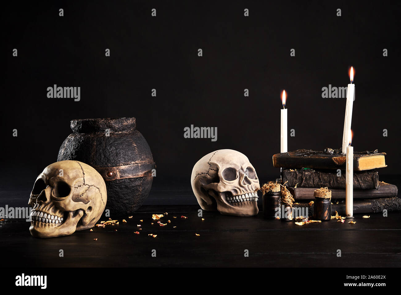 Realistic model of two craniums with white teeth, books with spells, jars of potion, old pot, burning candles are on a wooden dark table. The petals o Stock Photo