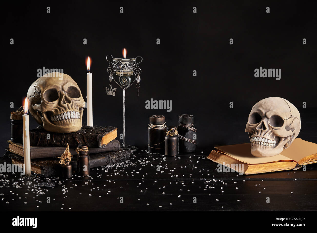 Realistic model of two human craniums with teeth are standing on an old books, jars, candlestick and burning candles are nearby on a wooden dark table Stock Photo