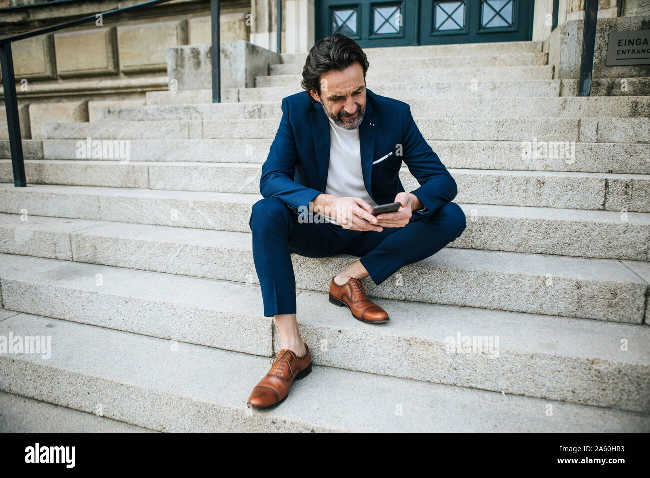 Bearded mature businessman wearing blue suit sitting on stairs looking at cell phone Stock Photo