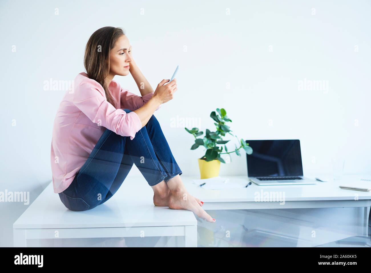 Young woman sitting on desk in office and using smartphone Stock Photo