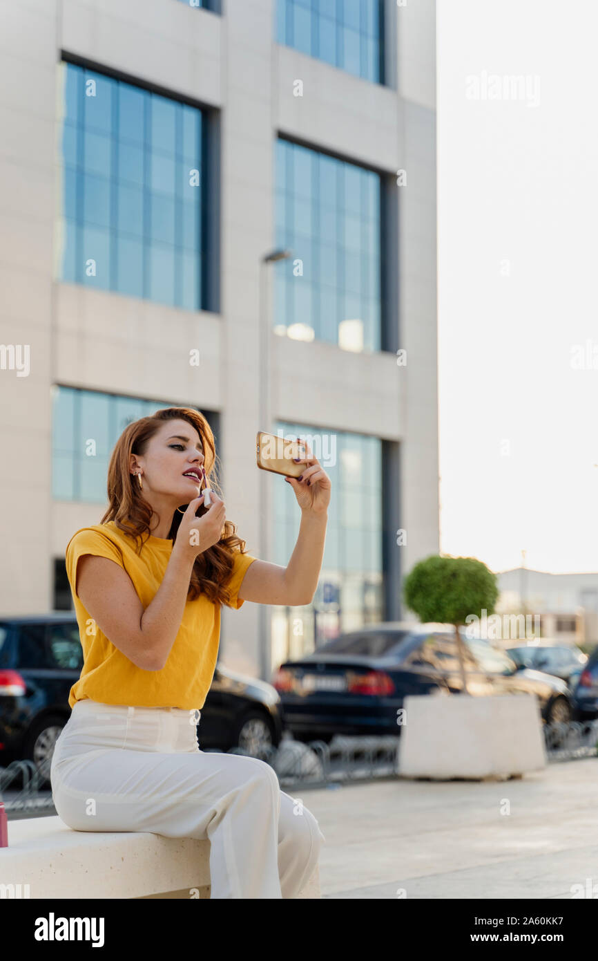 Businesswoman sitting in the sity, using smartphone to put on lipstick Stock Photo