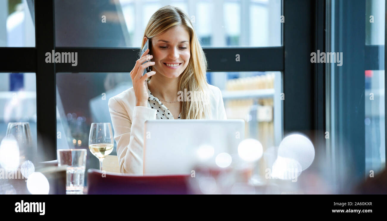Smiling businesswoman using cell phone and laptop in a restaurant Stock Photo