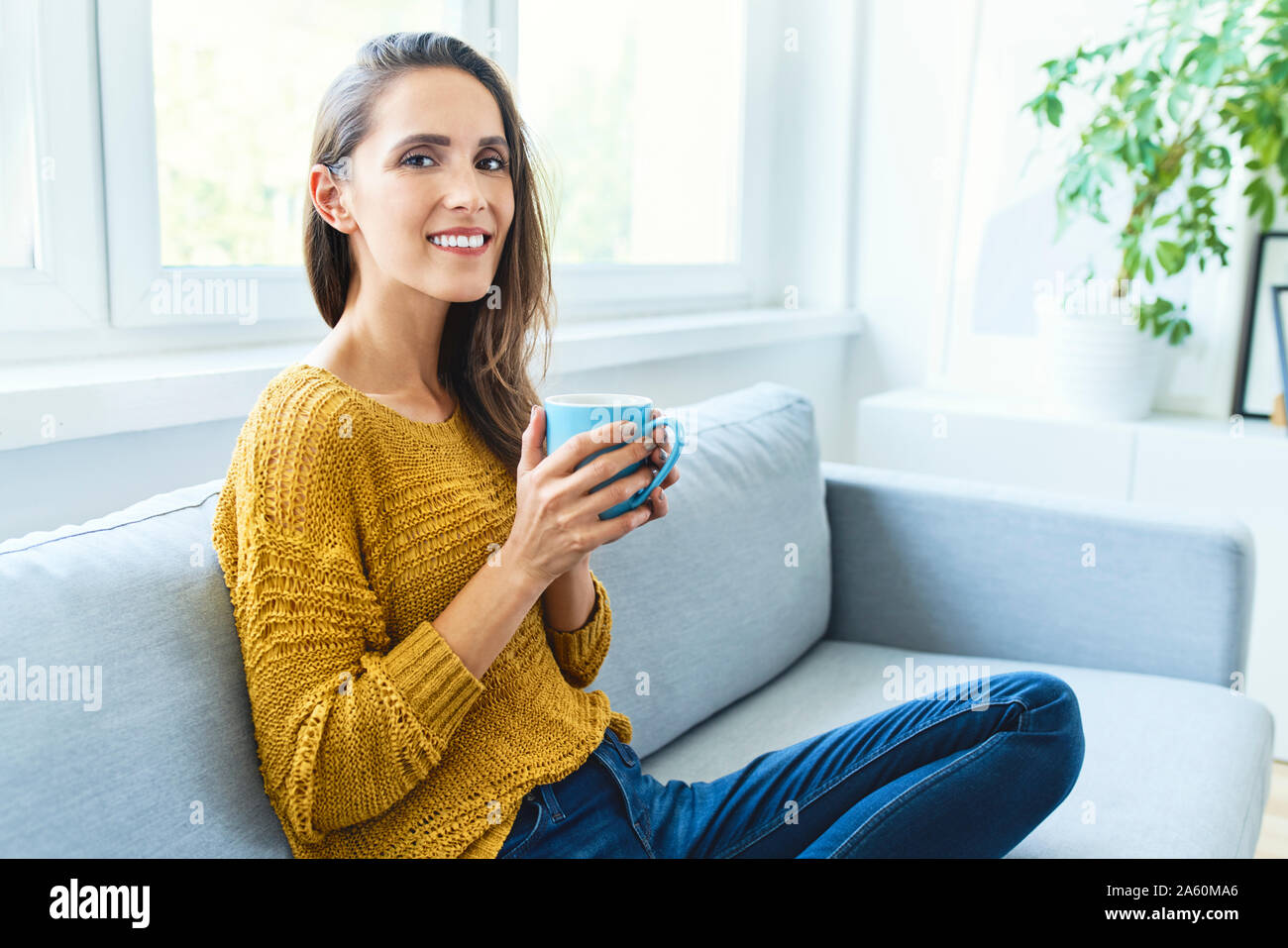 Portrait of young woman drinking coffee sitting on sofa and smiling at camera Stock Photo