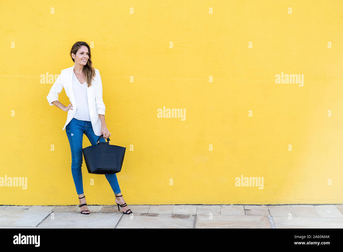 Smiling woman standing at a yellow wall holding a handbag Stock Photo