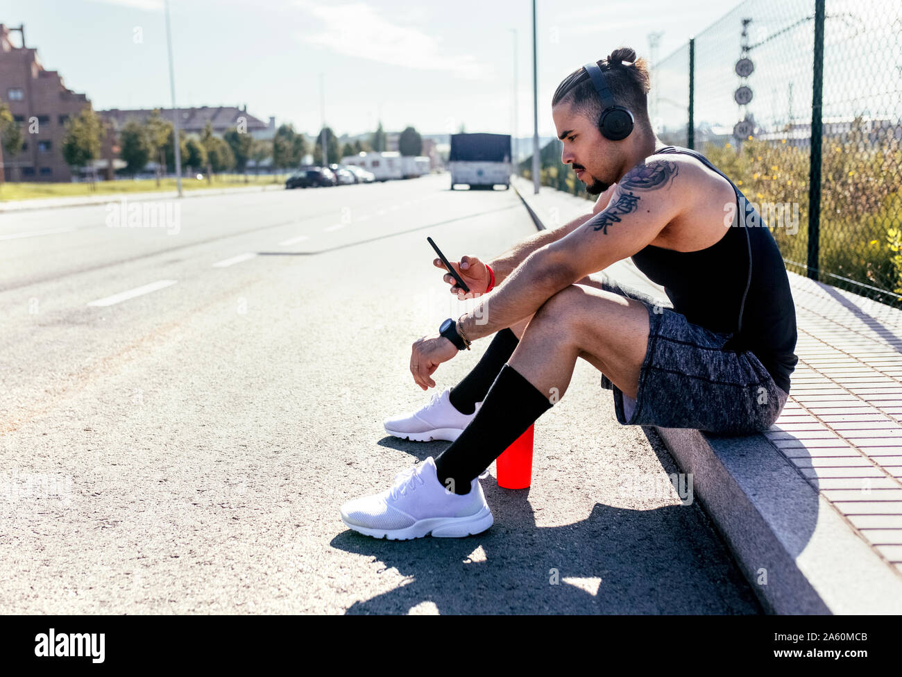 Sporty young man with smartphone and headphones sitting at the roadside Stock Photo