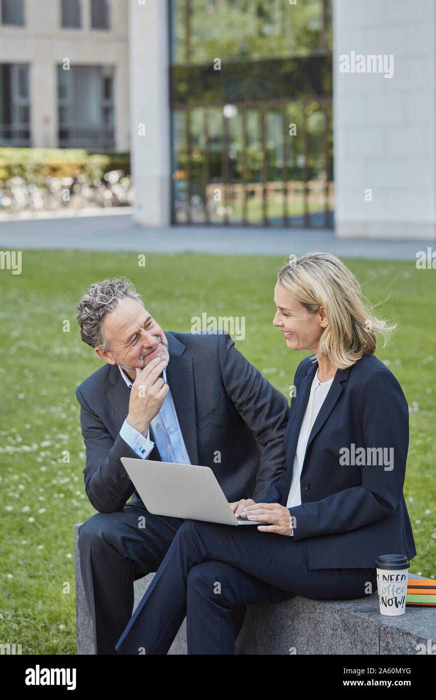 Businesswoman and businessman sitting on a wall in the city using laptop Stock Photo