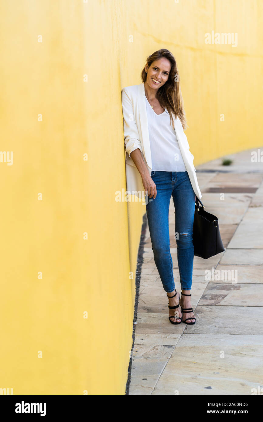 Portrait of smiling woman standing at a yellow wall holding a handbag Stock Photo