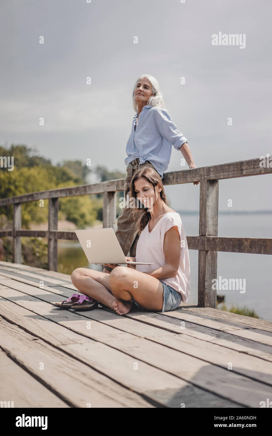 Woman sitting on wood bridge, using laptop, mother standing in background Stock Photo