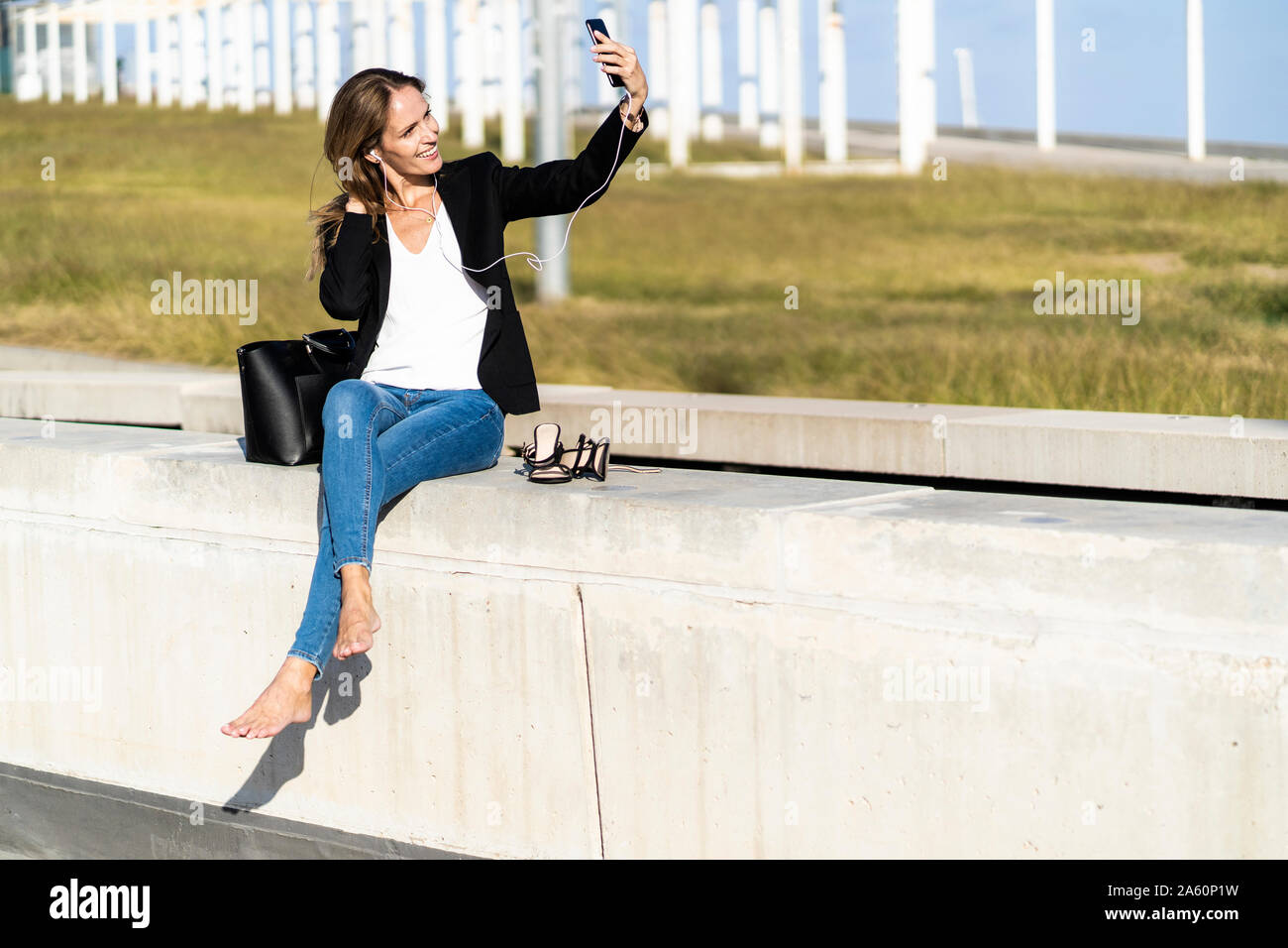 Smiling businesswoman sitting on a wall with smartphone and earphones Stock Photo