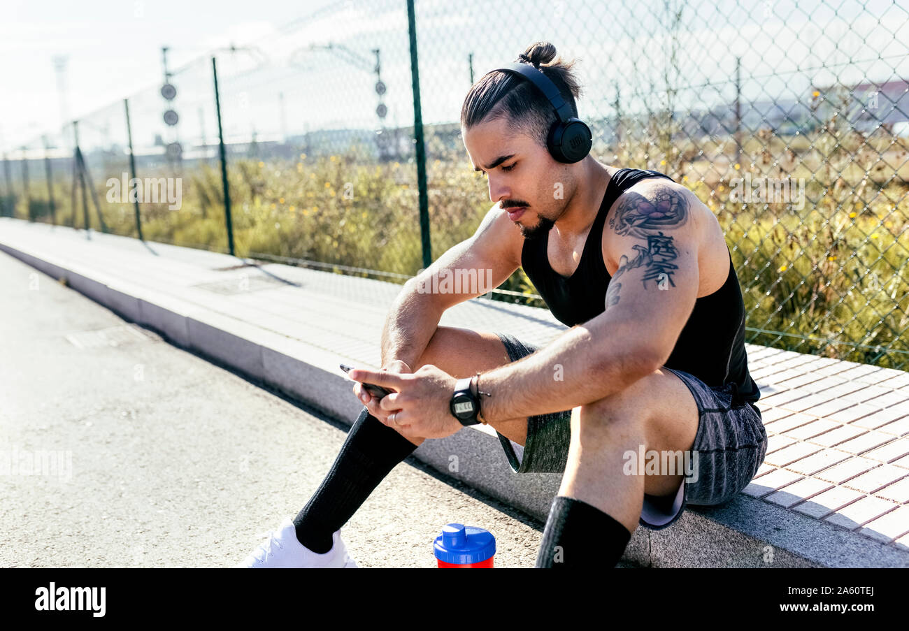 Sporty young man with smartphone and headphones sitting at the roadside Stock Photo