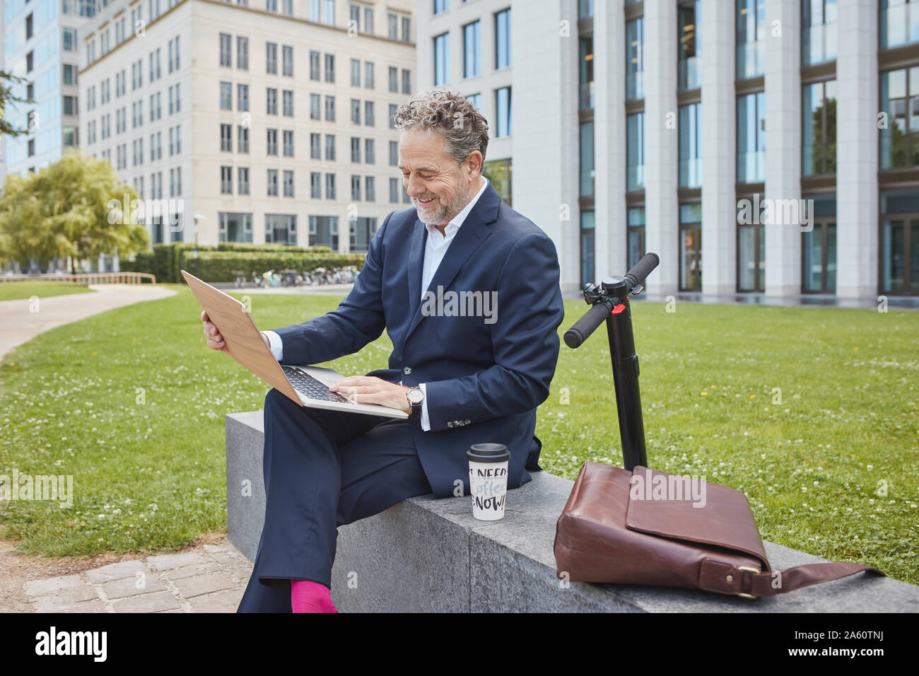 Happy mature businessman sitting on a wall in the city using laptop Stock Photo