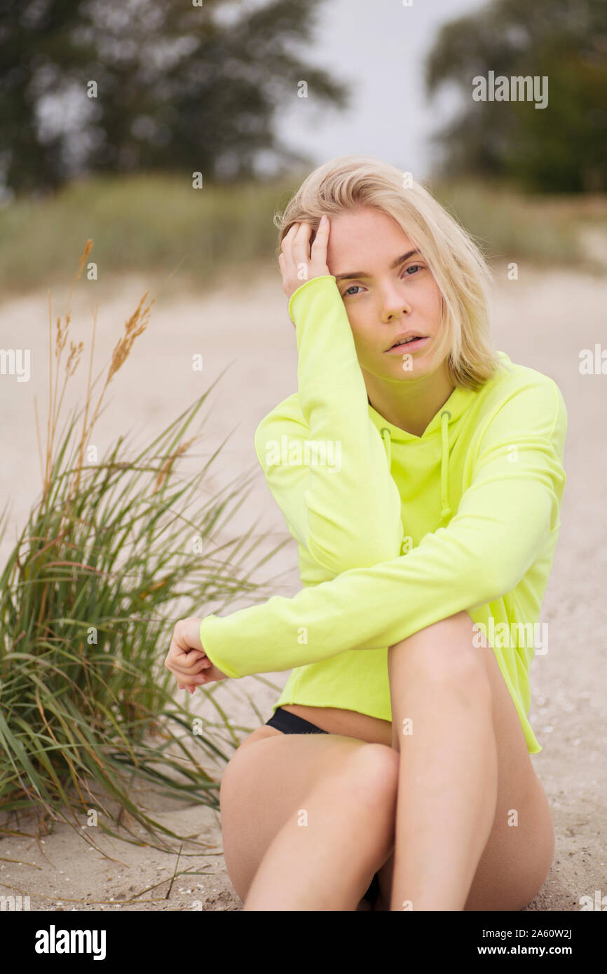 Portrait of blond young woman sitting on the beach wearing neon yellow sweatshirt Stock Photo