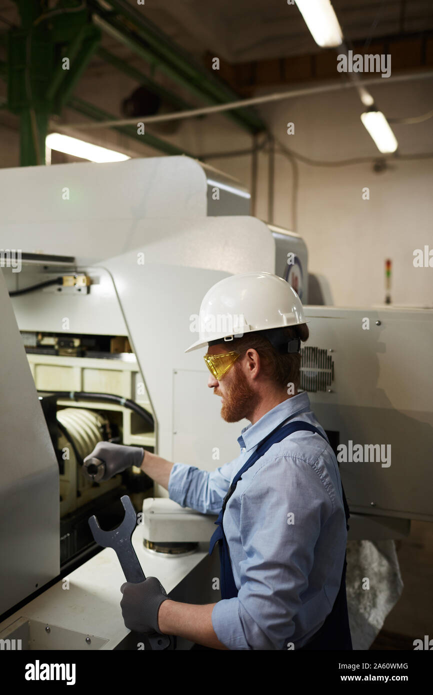 Bearded young man in work wear and protective glasses holding wrench and repairing the lathe Stock Photo