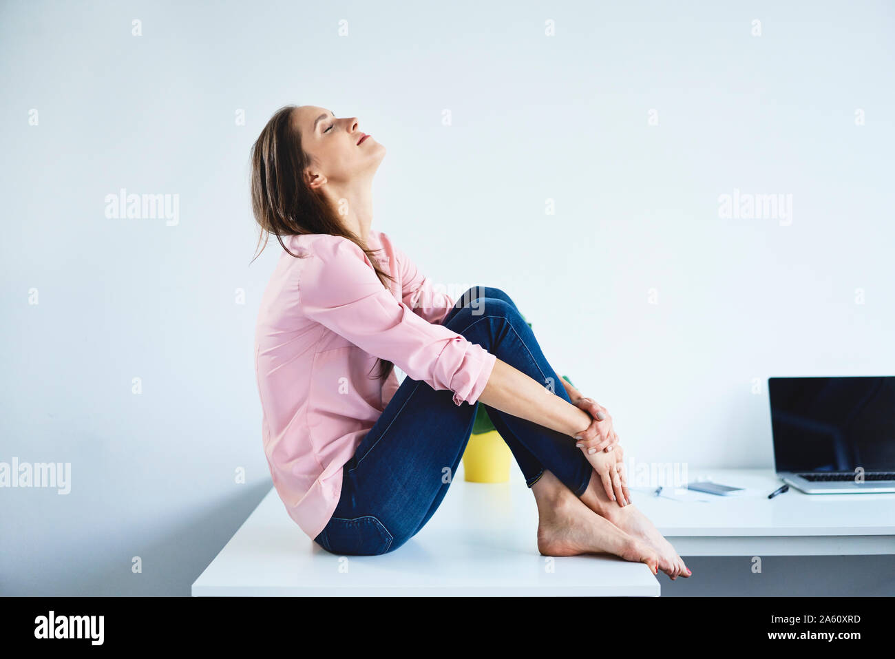 Young woman sitting on desk in office with eyes closed and relaxing Stock Photo