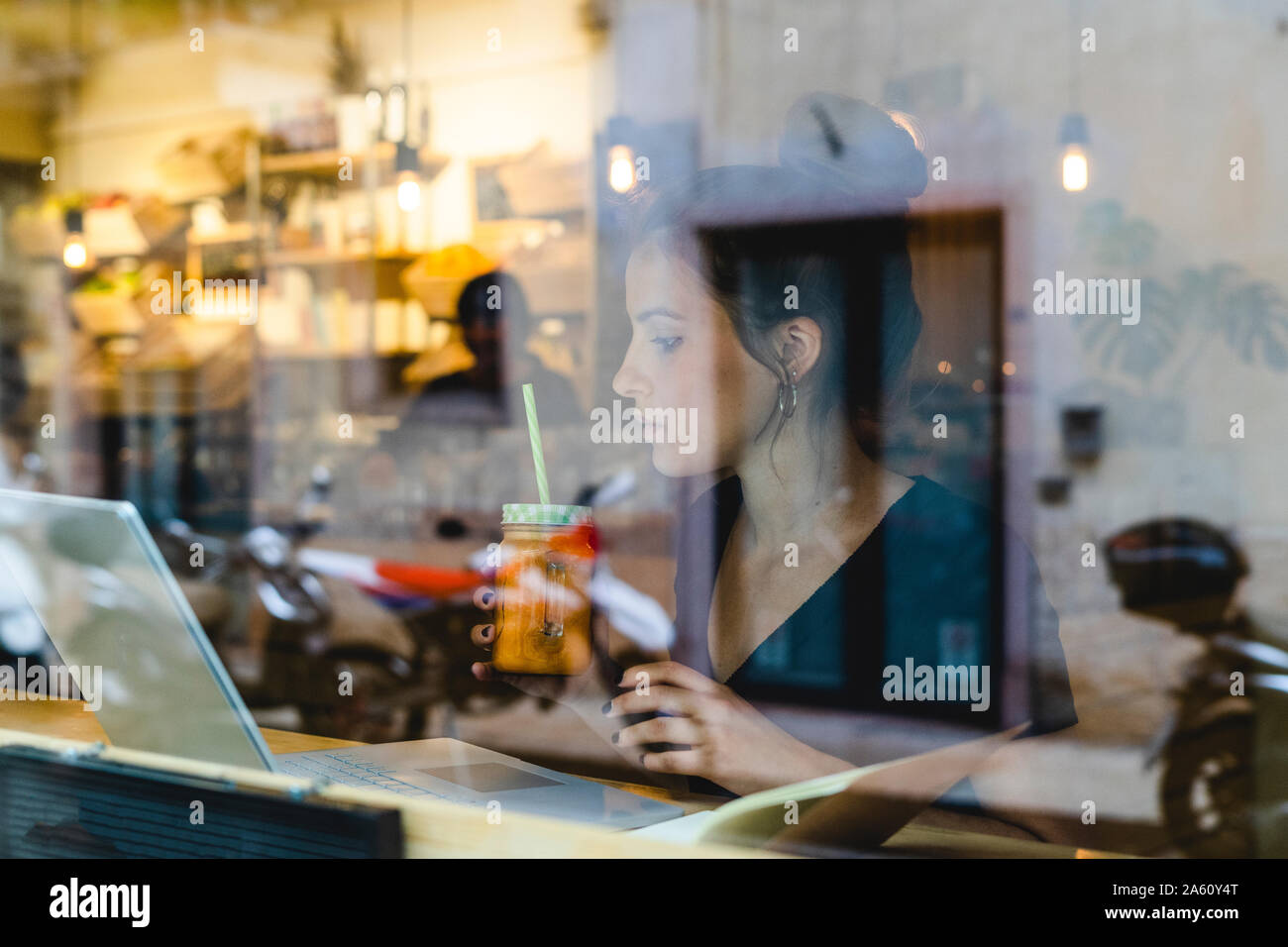 Young woman with smoothie and laptop behind windowpane in a cafe Stock Photo