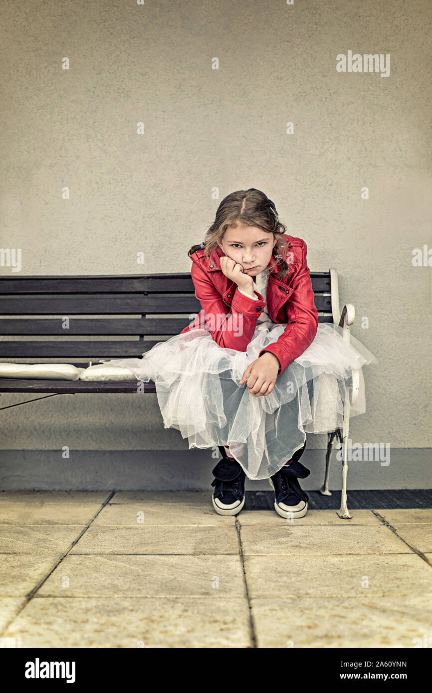Portrait of unhappy girl wearing red leather jacket and tutu sitting on bench Stock Photo