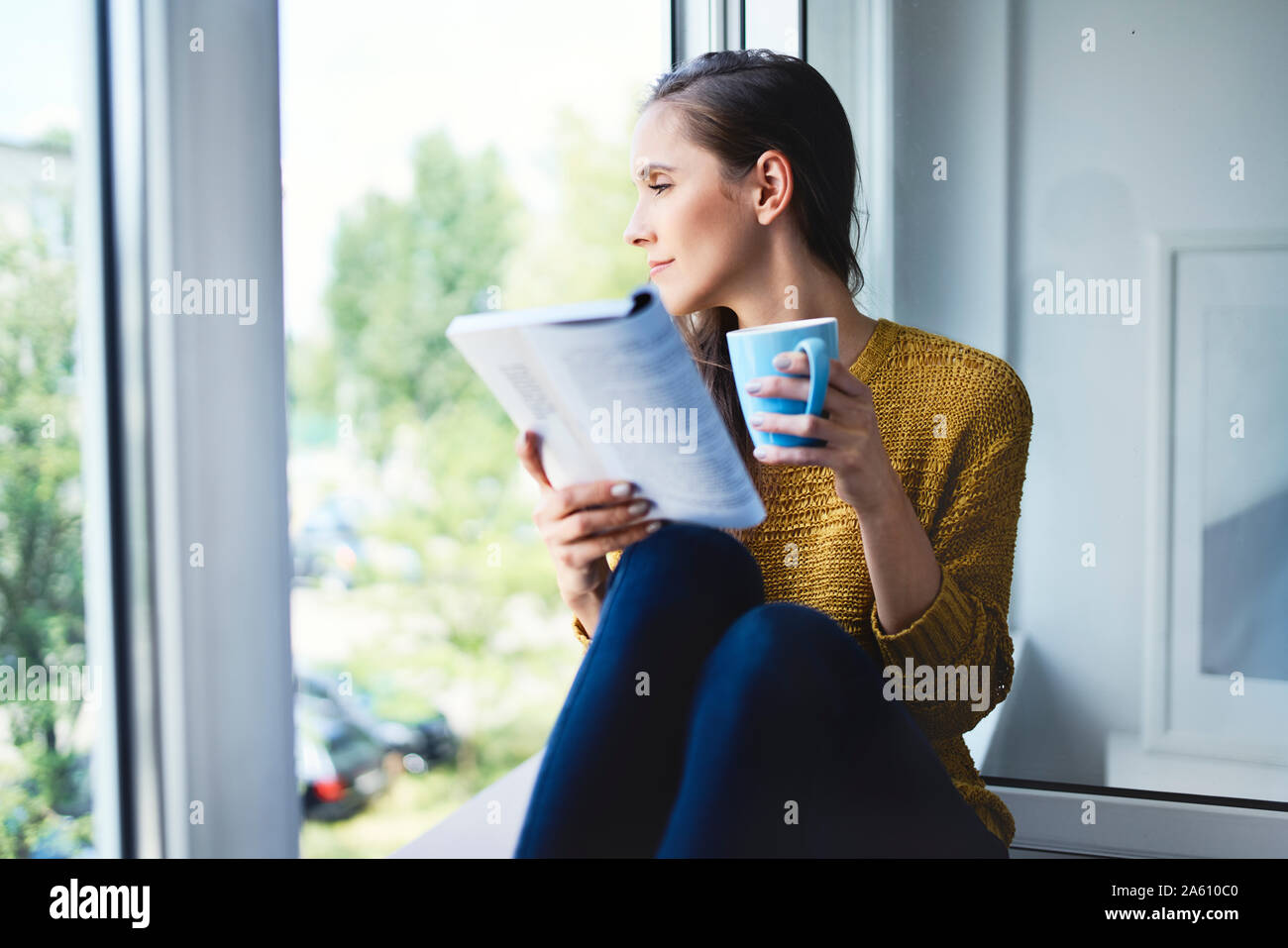 Young woman looking out the window while reading book at home Stock Photo