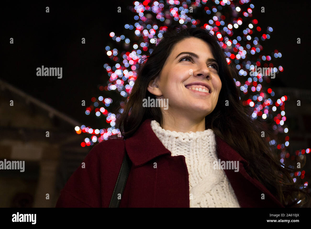 Portrait of happy young woman in front of lighted Christmas tree outdoors Stock Photo