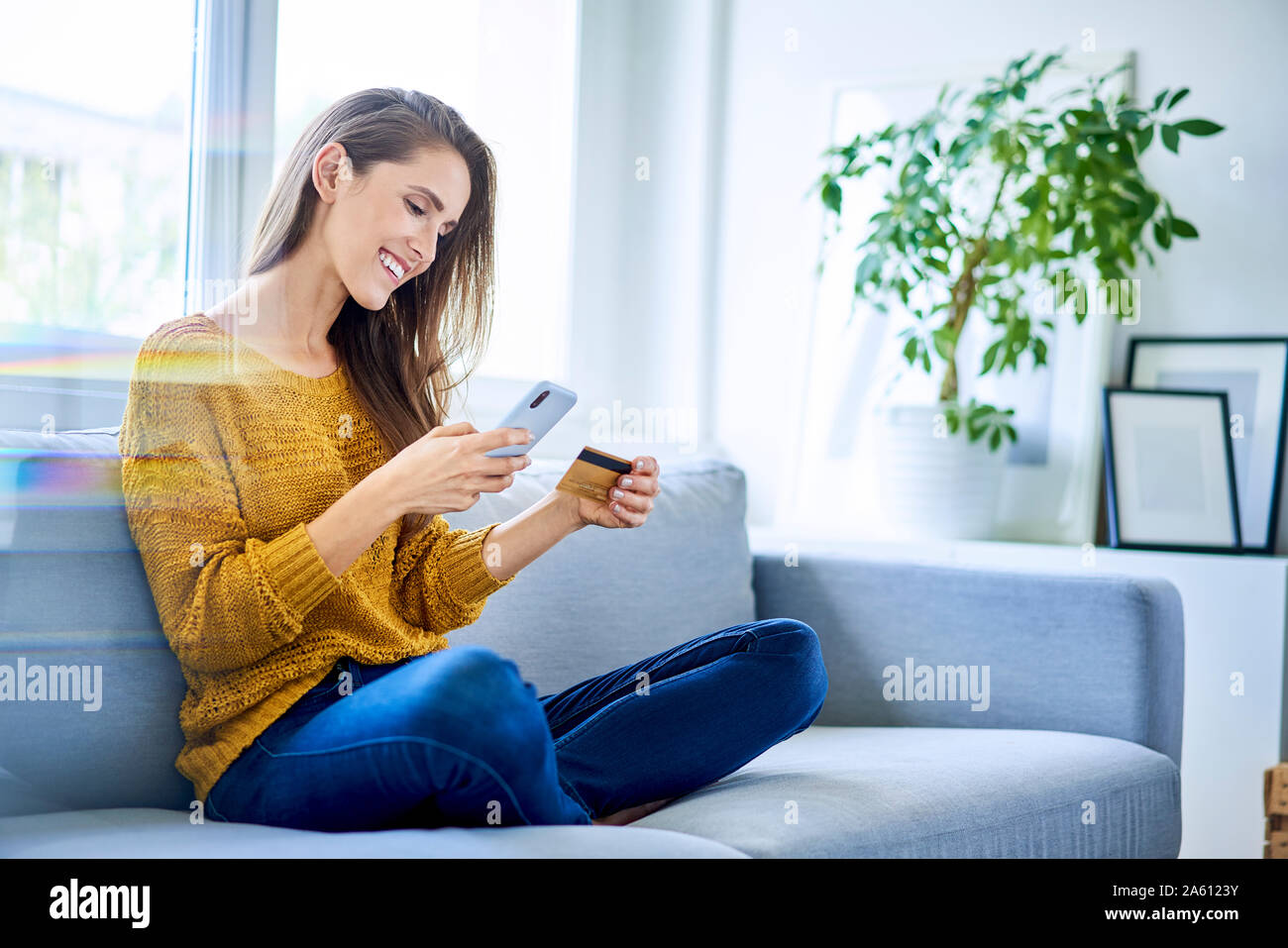 Beautiful happy woman paying online with smartphone and credit card while sitting on sofa Stock Photo