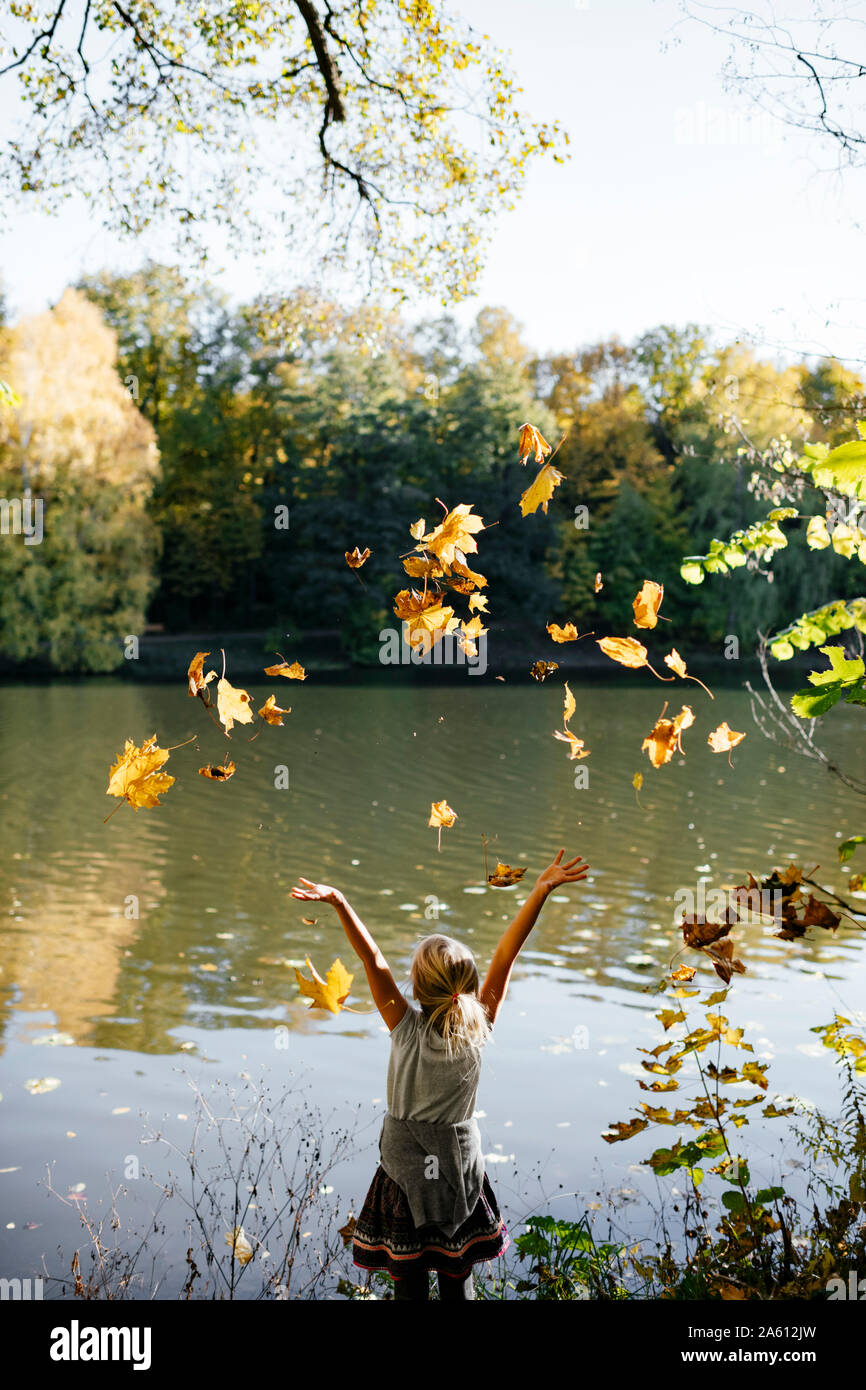 Back view of little girl standing at riverside throwing autumn leaves in the air Stock Photo