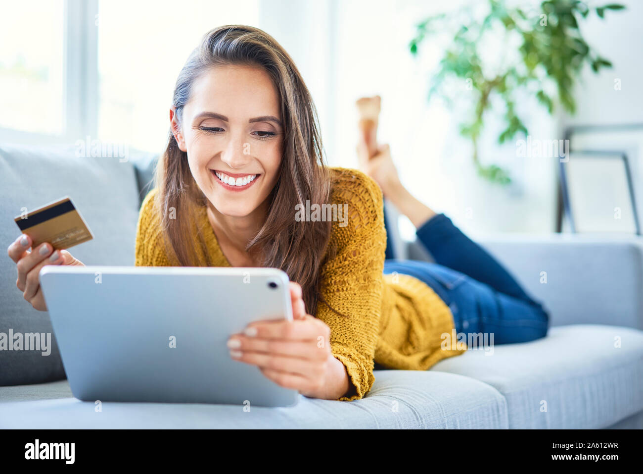 Cheerful young woman using credit card and tablet to shop online from home Stock Photo