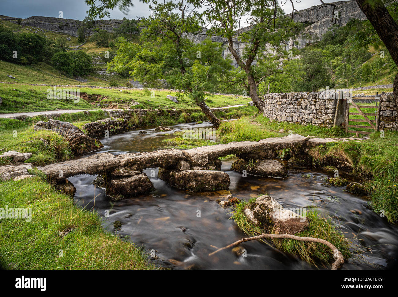 Old stone clapper bridge over Malham Beck with Malham Cove in the background Yorkshire Dales, England, UK. Stock Photo