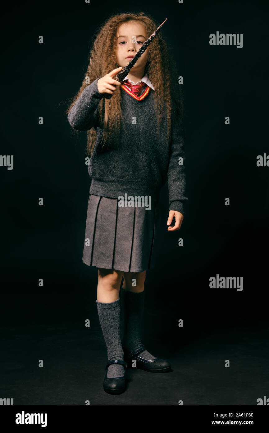 Portrait of a beautiful little witch girl with magnificent long brown hair dressed in a navy blue jumper, gray skirt, knee socks and shoes. She is hol Stock Photo