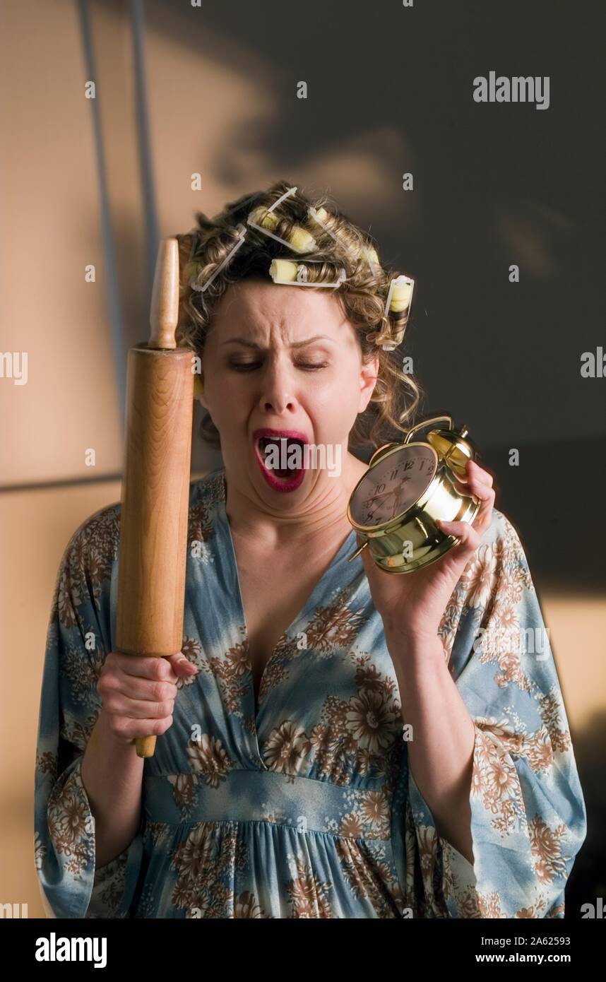 A humorous photo of a yawning woman in hair curlers holding a rolling pin and alarm clock, waiting for her man to come home Stock Photo