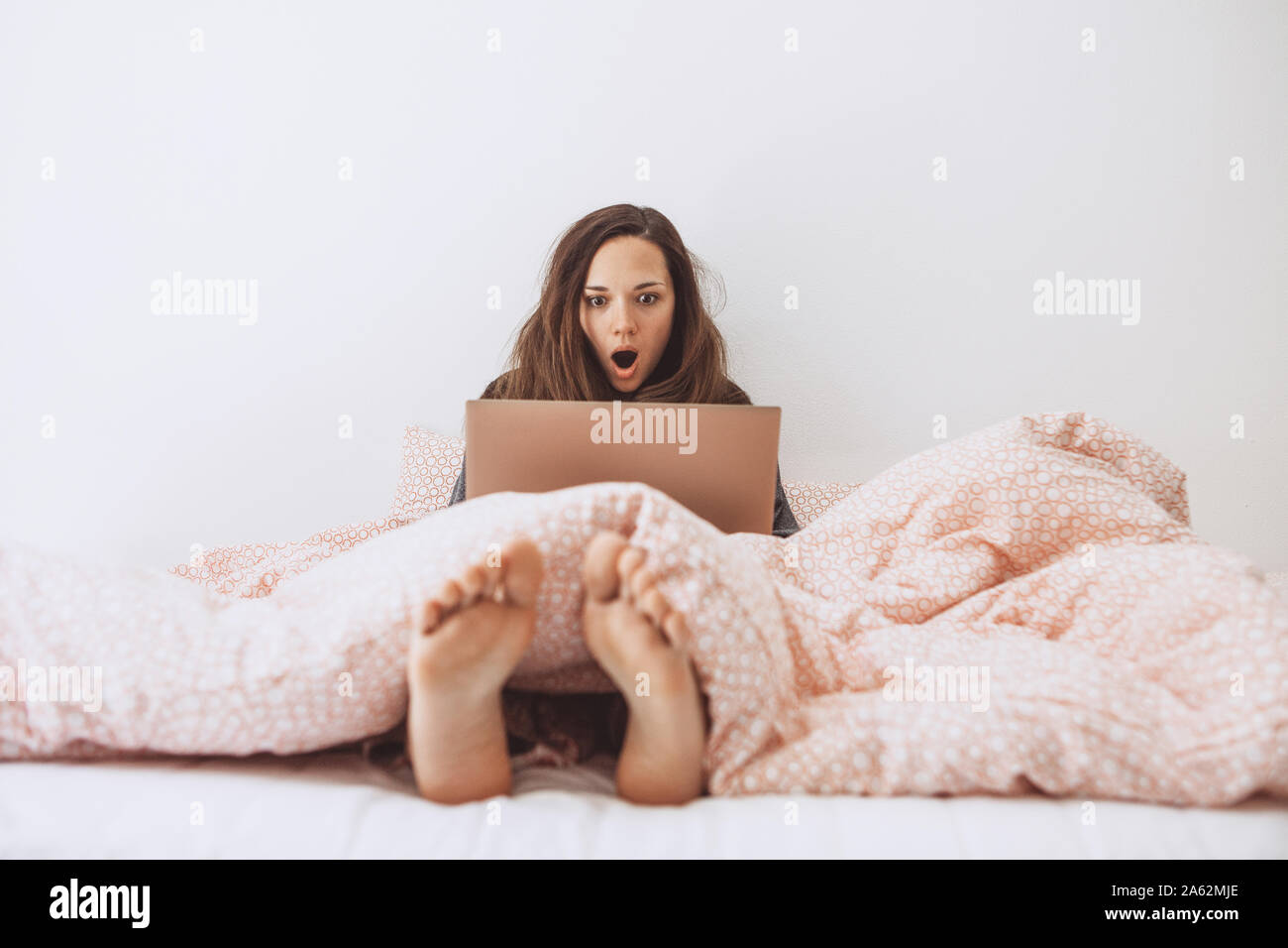The girl uses the computer in bed. She saw something and was surprised or shocked. Stock Photo