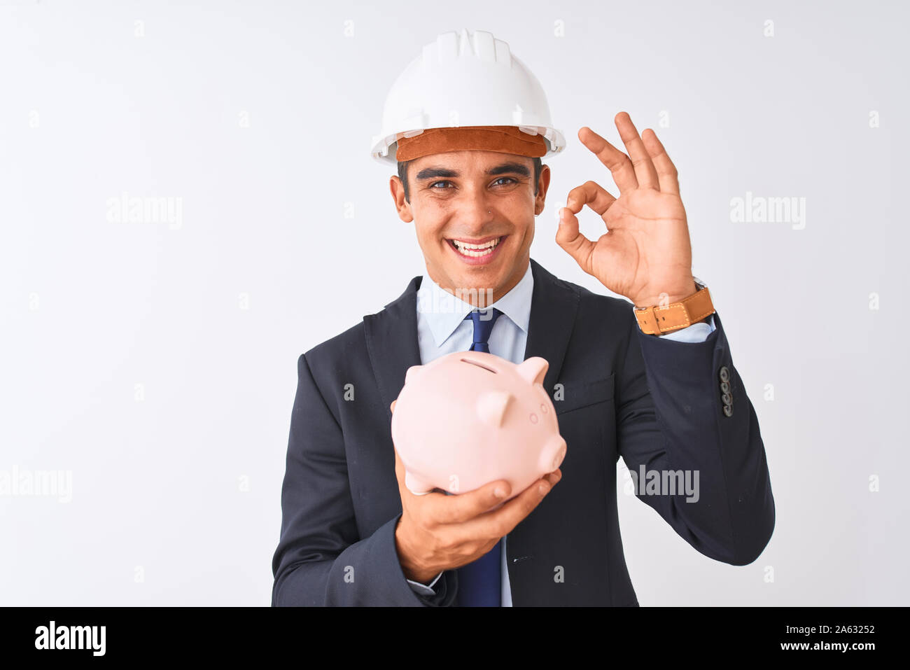 Handsome architect man wearing helmet holding piggy bank over isolated ...