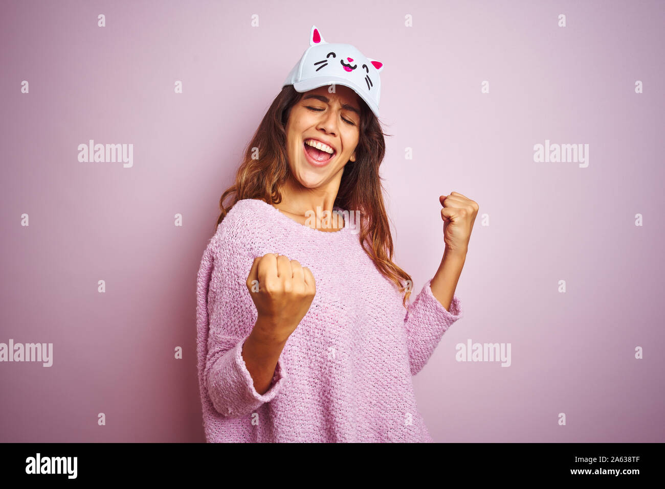 Young beautiful woman wearing funny cat cap standing over pink isolated background very happy and excited doing winner gesture with arms raised, smili Stock Photo
