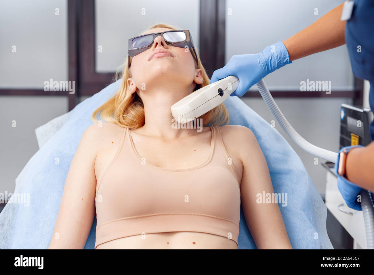 Cosmetology Service. Young woman at beauty clinic lying on medical bed in safety goggles while doctor using photorejuvenation laser on her neck Stock Photo