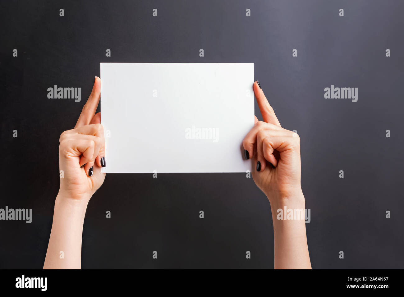 Woman's hands with black nailpolish holding a blank paper against black wall Stock Photo