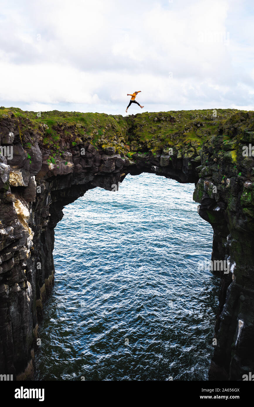 Tourist jumps over a natural rock bridge in Arnarstapi, Iceland Stock Photo