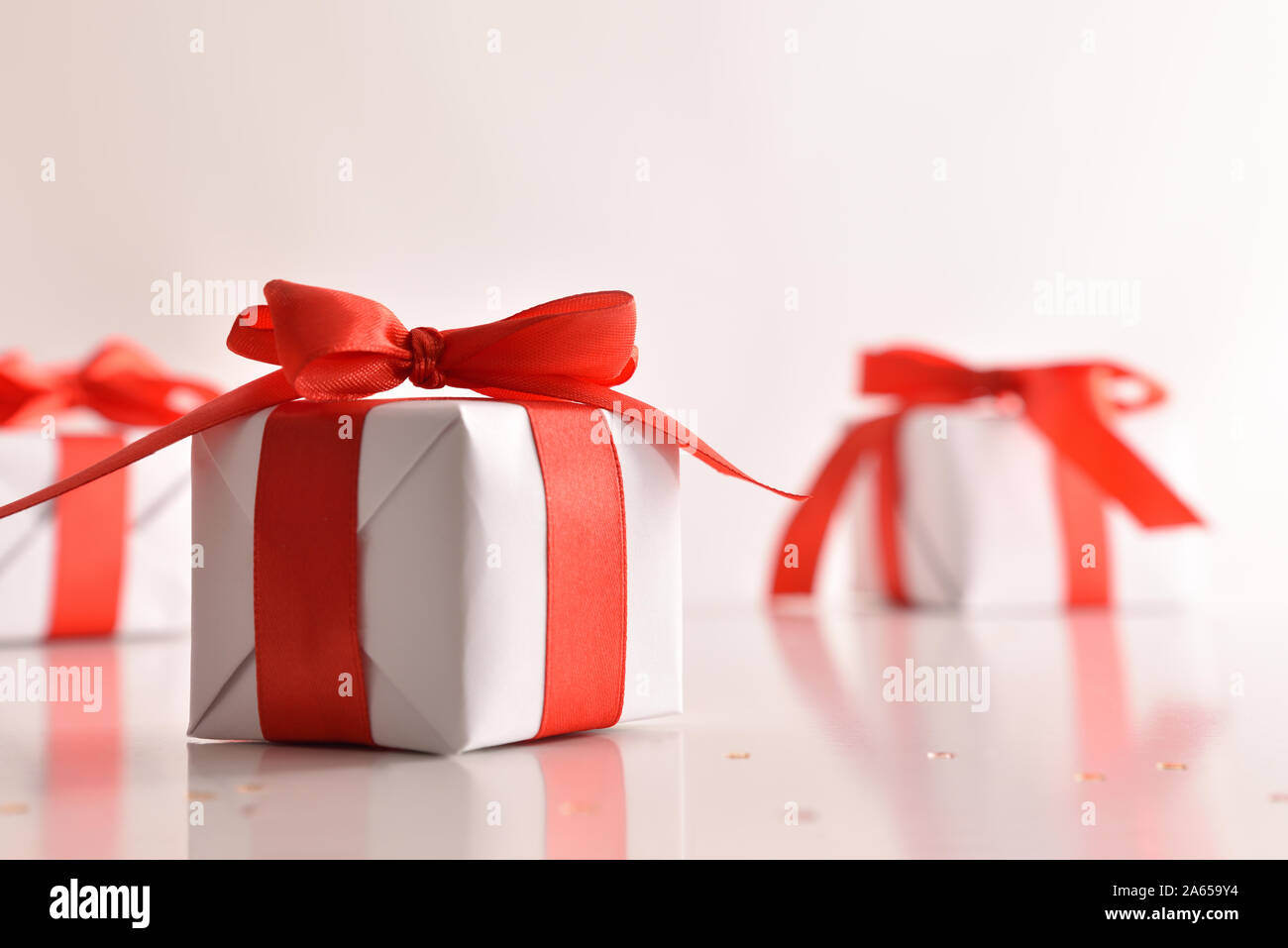 Three white paper gift boxes with red bow on white reflective table and background.. Horizontal composition. Front view. Stock Photo