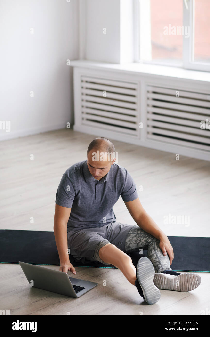 Young disabled man sitting on the floor on exercise mat and watching online sports training on laptop in the room Stock Photo