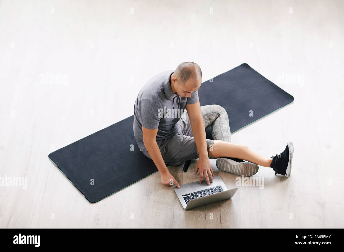 Young disabled man sitting on exercise mat and communicating online using his laptop computer in the room Stock Photo