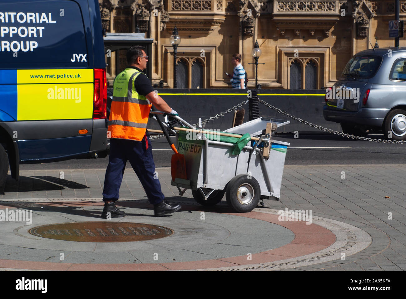 A road sweeper, street cleaner, pushing his trolley past the Houses of Parliament, Westminster,London, wearing a hi-viz jacketand two vehicles in view Stock Photo