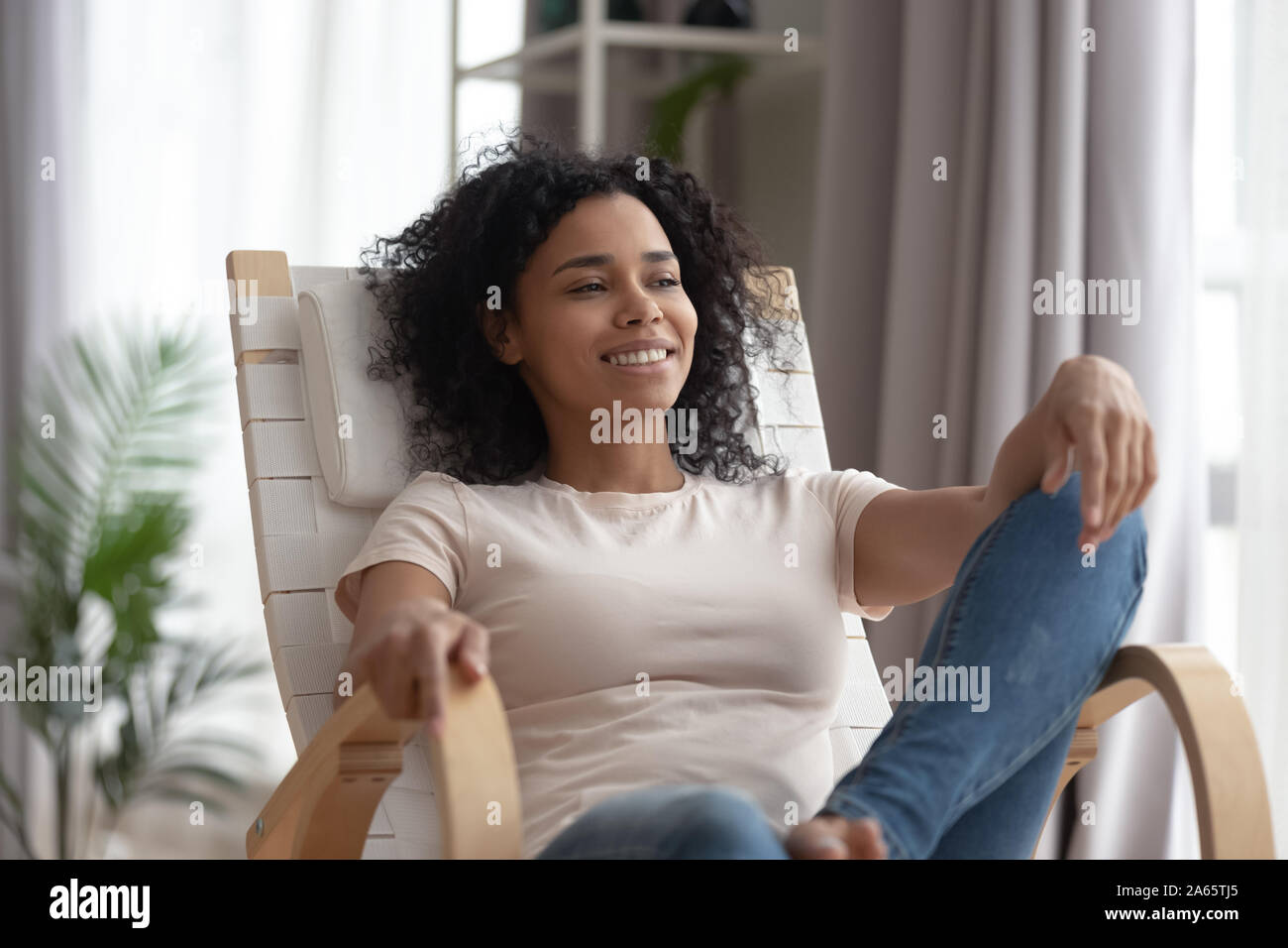 Smiling black young woman relax in chair looking in distance Stock Photo