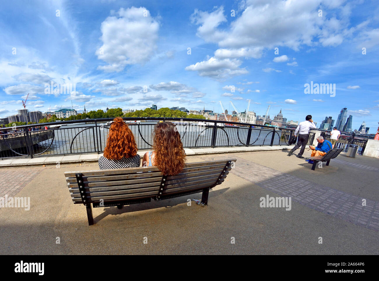 London, England, UK. Two young women sitting on a bench on the South Bank, looking over the River Thames Stock Photo