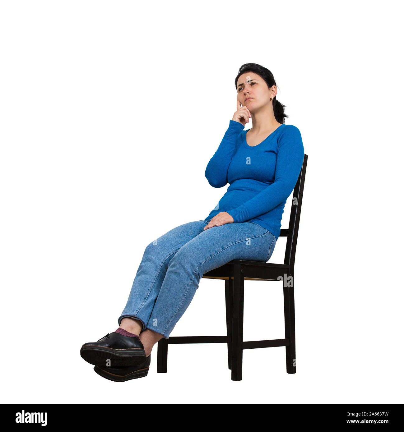 Side view full length portrait of casual young woman seated on a chair keeps hand under chin thoughtful looking away isolated over white background wi Stock Photo